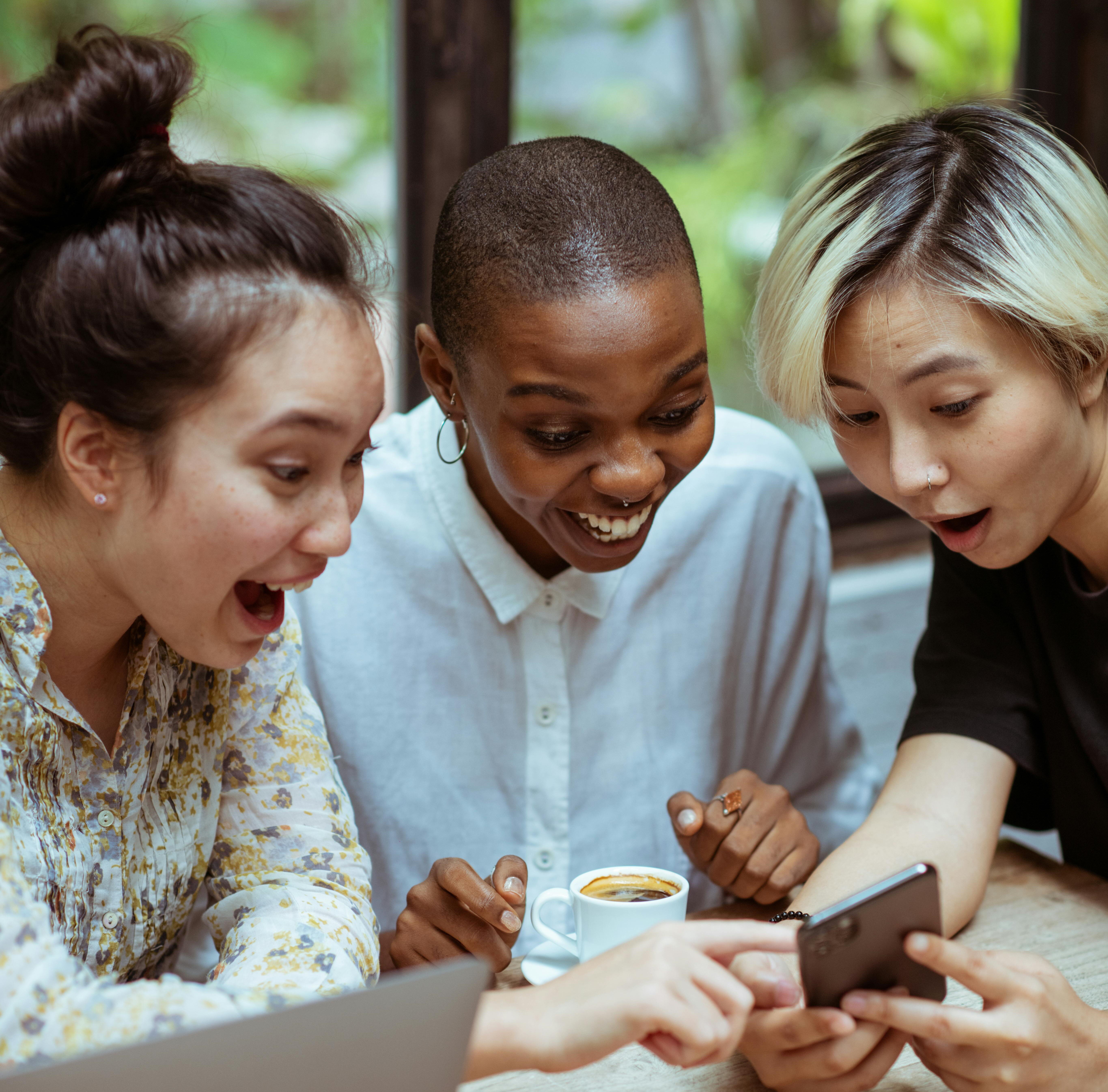 tres chicas de diversidad étnica rodean en una mesa a un celular y estan sonrientes y sorprendidas de ver el mensaje